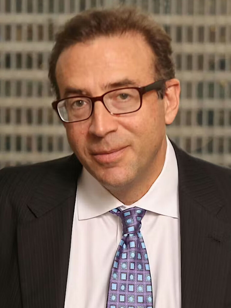 Michael Cembalest. A man with short brown hair and glasses, wearing a dark pinstripe suit, white shirt, and patterned blue tie, sits in front of a window with a blurred cityscape background at the Healthcare Leadership Exchange.