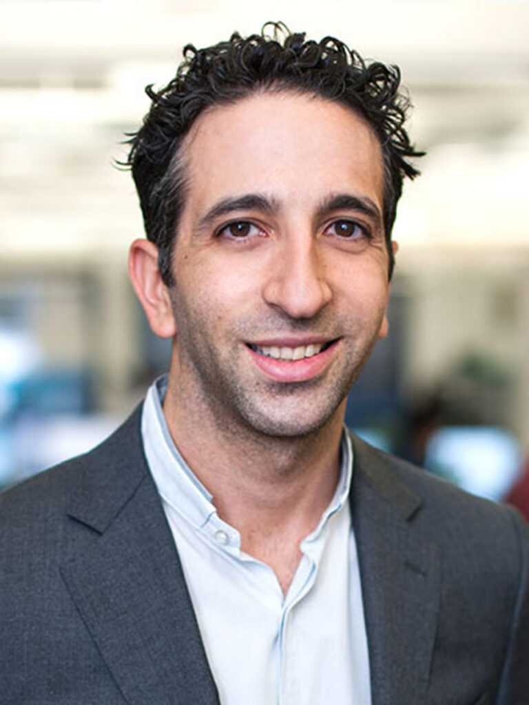 Reed Mollins. A man with short, curly dark hair wearing a gray suit jacket and a light-colored shirt smiles at the camera in an office setting, representing the spirit of collaboration at the Healthcare Leadership Exchange.