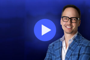 A smiling man in glasses and a blue plaid jacket stands in front of a blue background with a large white play button icon in the center.