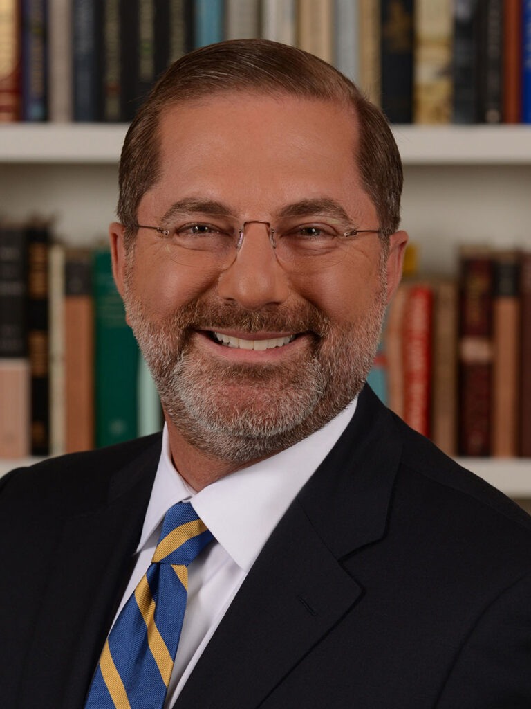 Alex Azar in a suit and striped tie smiles in front of a bookshelf filled with books, embodying the spirit of the Healthcare Leadership Exchange.