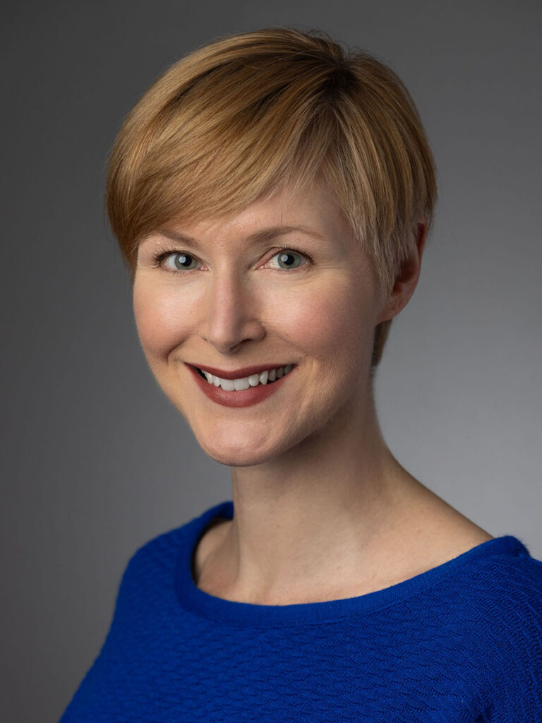 Molly Smith. A woman with short blond hair wearing a blue textured top, smiling at the camera against a plain gray background, represents the spirit of connection fostered by Healthcare Leadership Exchange.