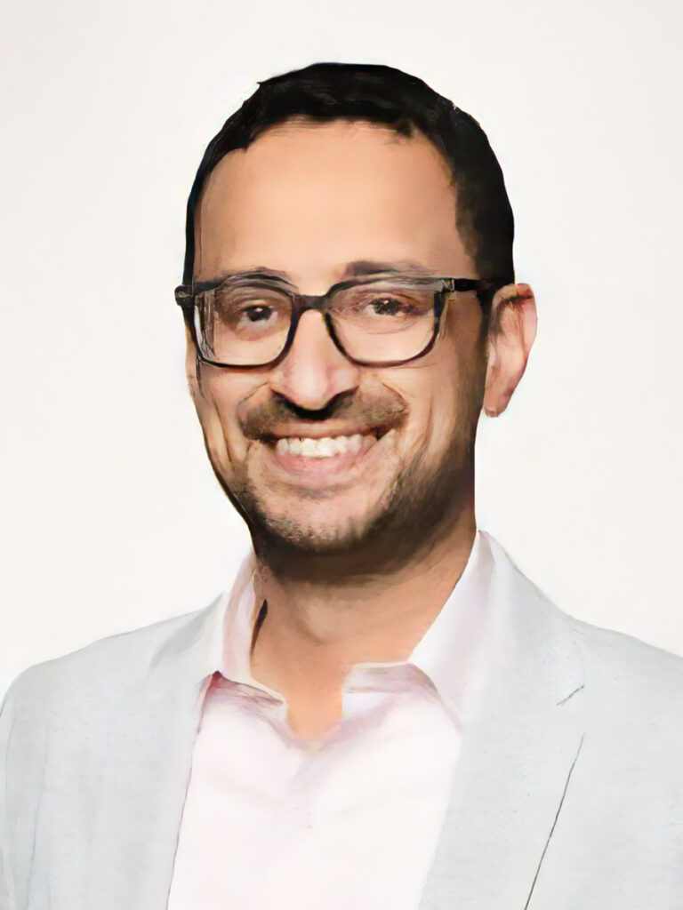 Syed Mohiuddin wearing glasses and a light-colored blazer over a light pink shirt smiles at the camera against a plain background, embodying professionalism seen at Healthcare Leadership Exchange events.