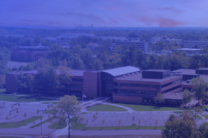 Aerial view of Ensemble. A large, modern building surrounded by trees and parking lots, with additional buildings and forested areas in the background. Image has a blue color overlay.