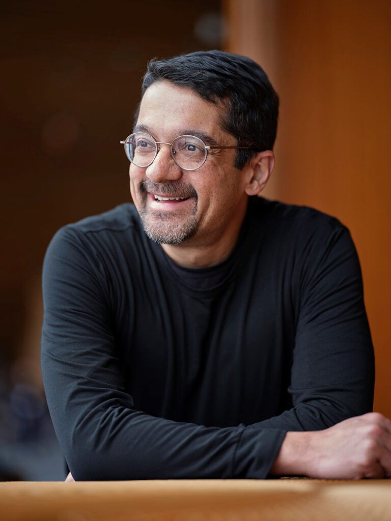 Shankar Vedantam, with short dark hair and glasses, wearing a black long-sleeve shirt, smiles while leaning on a surface with his arms crossed.