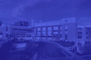 Modern hospital building with multiple floors, large windows, and a curved driveway in front, photographed during early evening with cloudy sky.