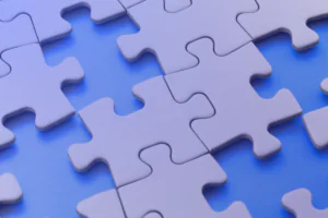 Several white puzzle pieces, representing denials management, lie on a blue surface—some connected and some separated—with soft lighting creating a slight purple tint.
