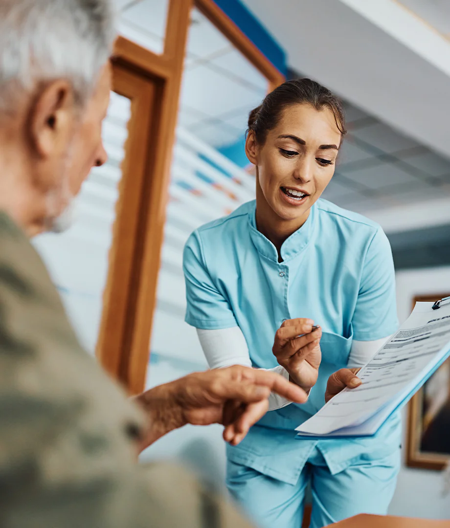 A nurse in blue scrubs shows a clipboard with documents to an older man, explaining information as they interact in a bright indoor setting.