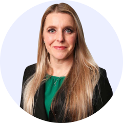 Stacie Sutter A woman with long blonde hair wearing a black blazer over a green top, standing against a plain light background, looking at the camera with a neutral expression, exuding the confidence of someone experienced in payer strategy.